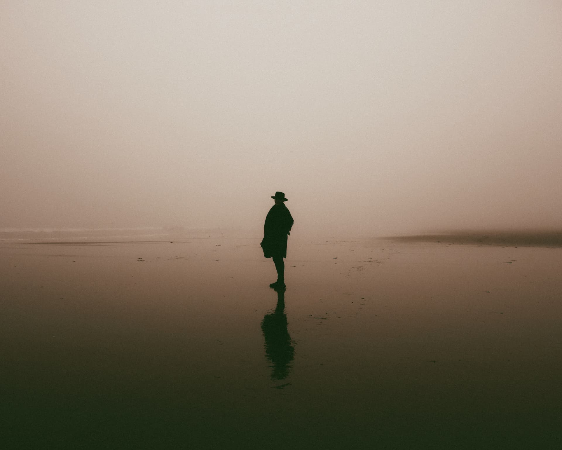 A person in a wide-brimmed hat reflected on wet sand in the mist