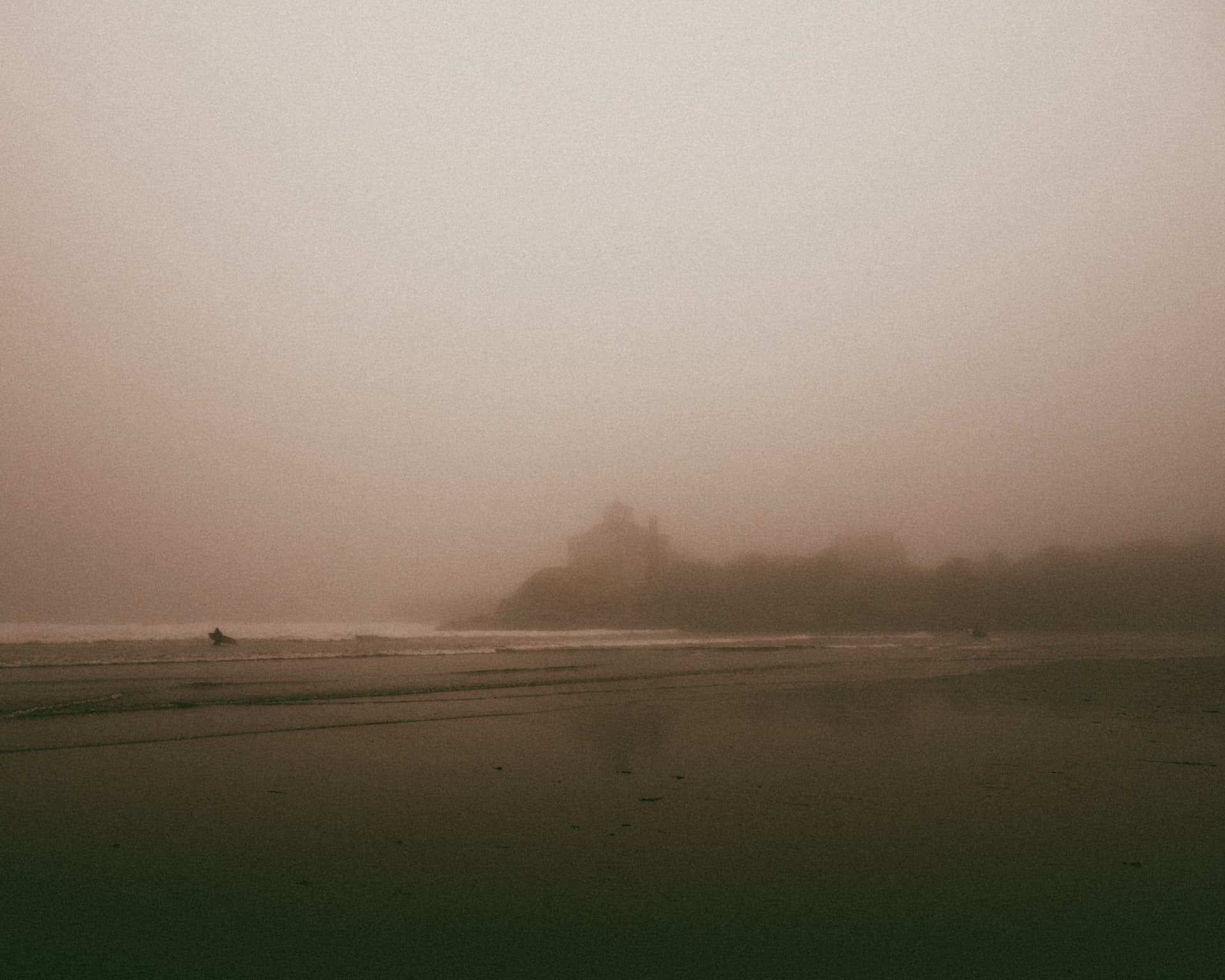 A misty beach with distant surfers and a headland fading into fog