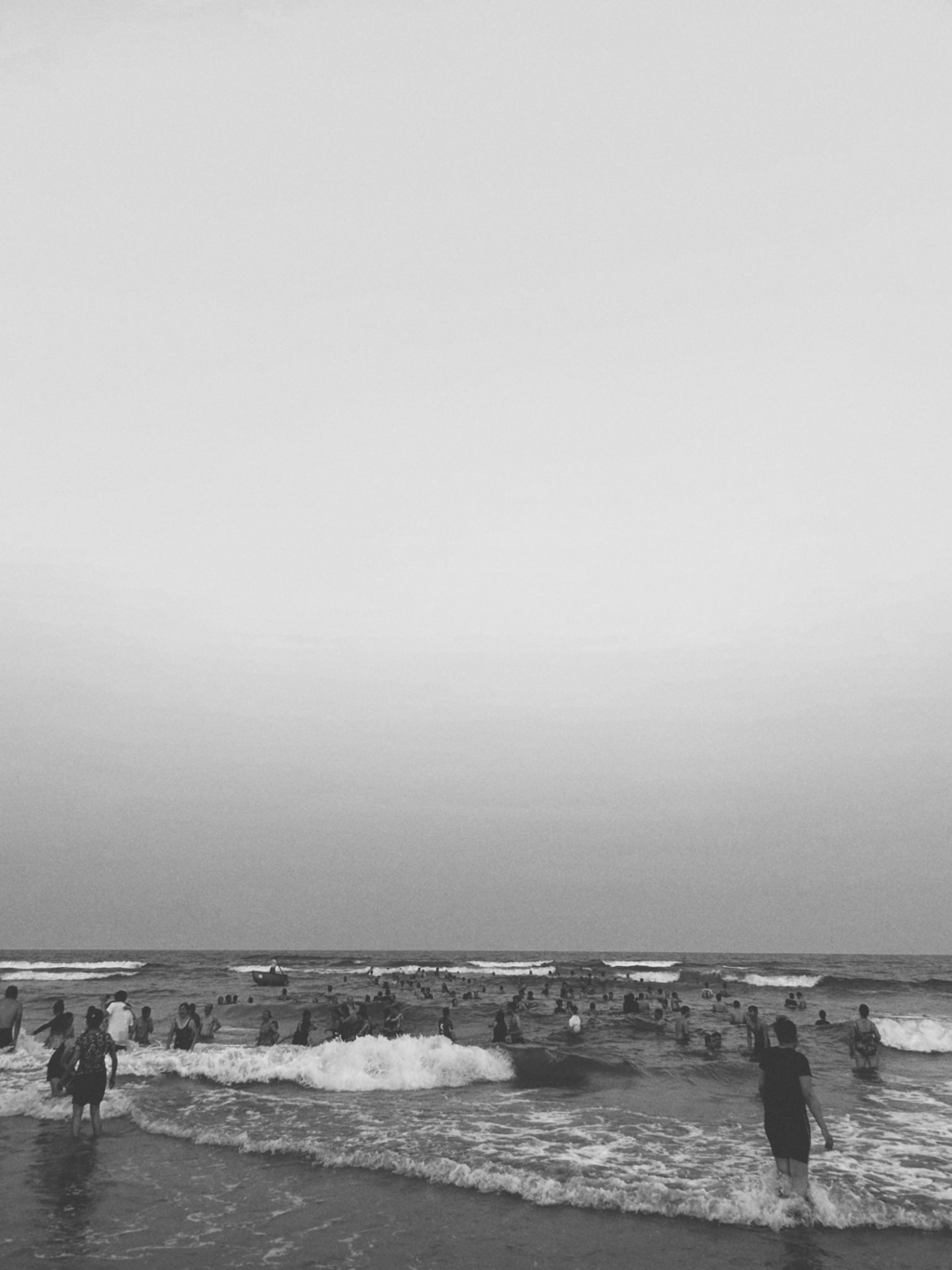 Crowds wading in calm surf beneath a pale sky