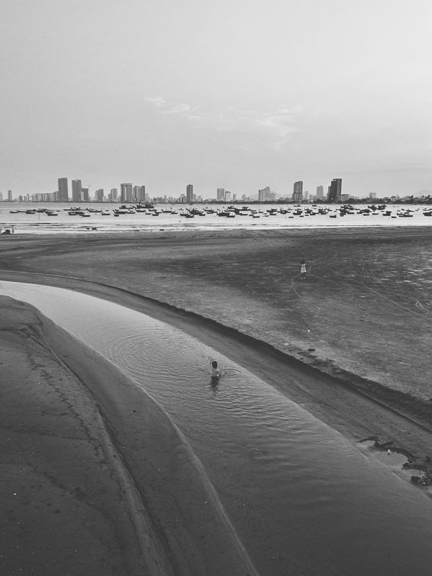 A child wading a tidal stream with a city rising behind the boats