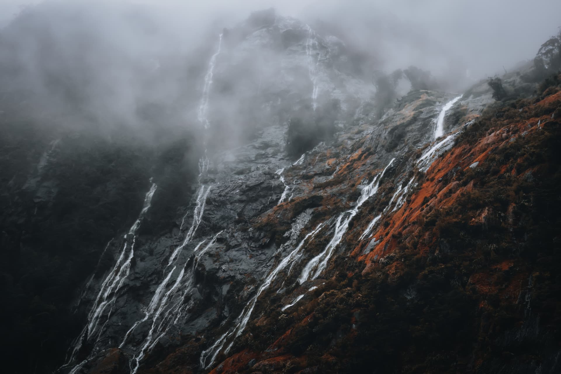 A waterfall-streaked mountainside veined with orange moss in fog